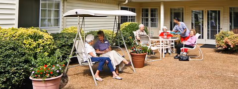Several elderly people sitting and chatting on an outdoor patio with a swing, chairs, tables, and potted plants in front of a building.