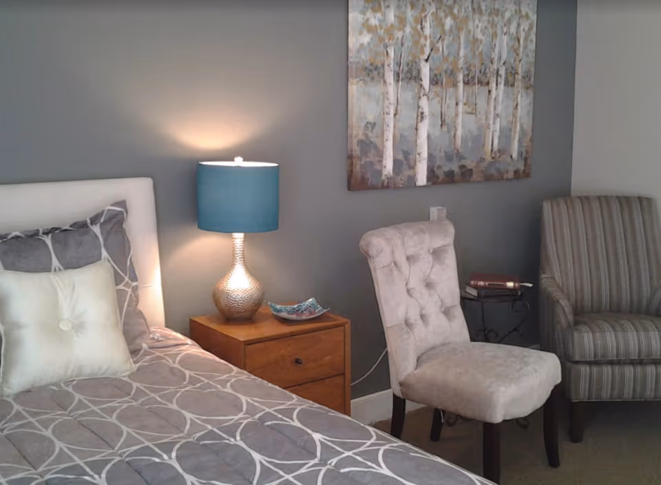 A cozy bedroom corner featuring a bed with patterned bedding and pillows, a wooden nightstand with a decorative lamp and a small dish, a tufted beige chair, a striped armchair, and a painting of birch trees on a gray wall.