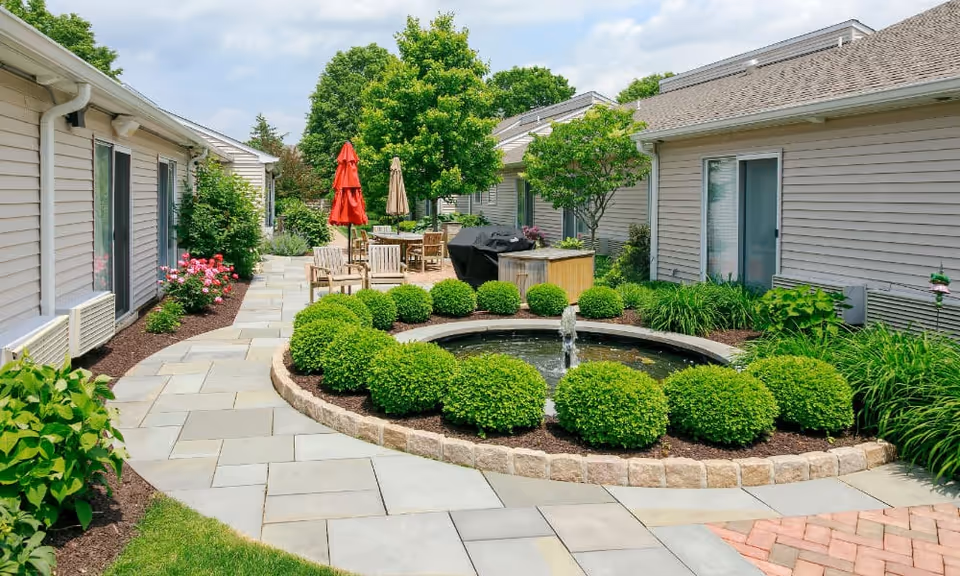 A landscaped outdoor courtyard with a circular fountain, trimmed shrubs, patio seating and umbrellas between single-story building facades.