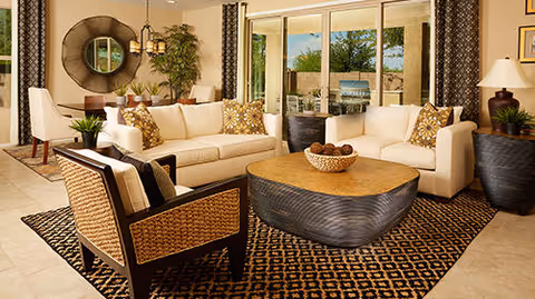 A spacious living room with two white sofas, a woven armchair, a large patterned rug, and a wooden coffee table with decorative items. The room has large windows with a view of an outdoor patio area, beige tiled floors, and warm lighting with a chandelier and table lamps.