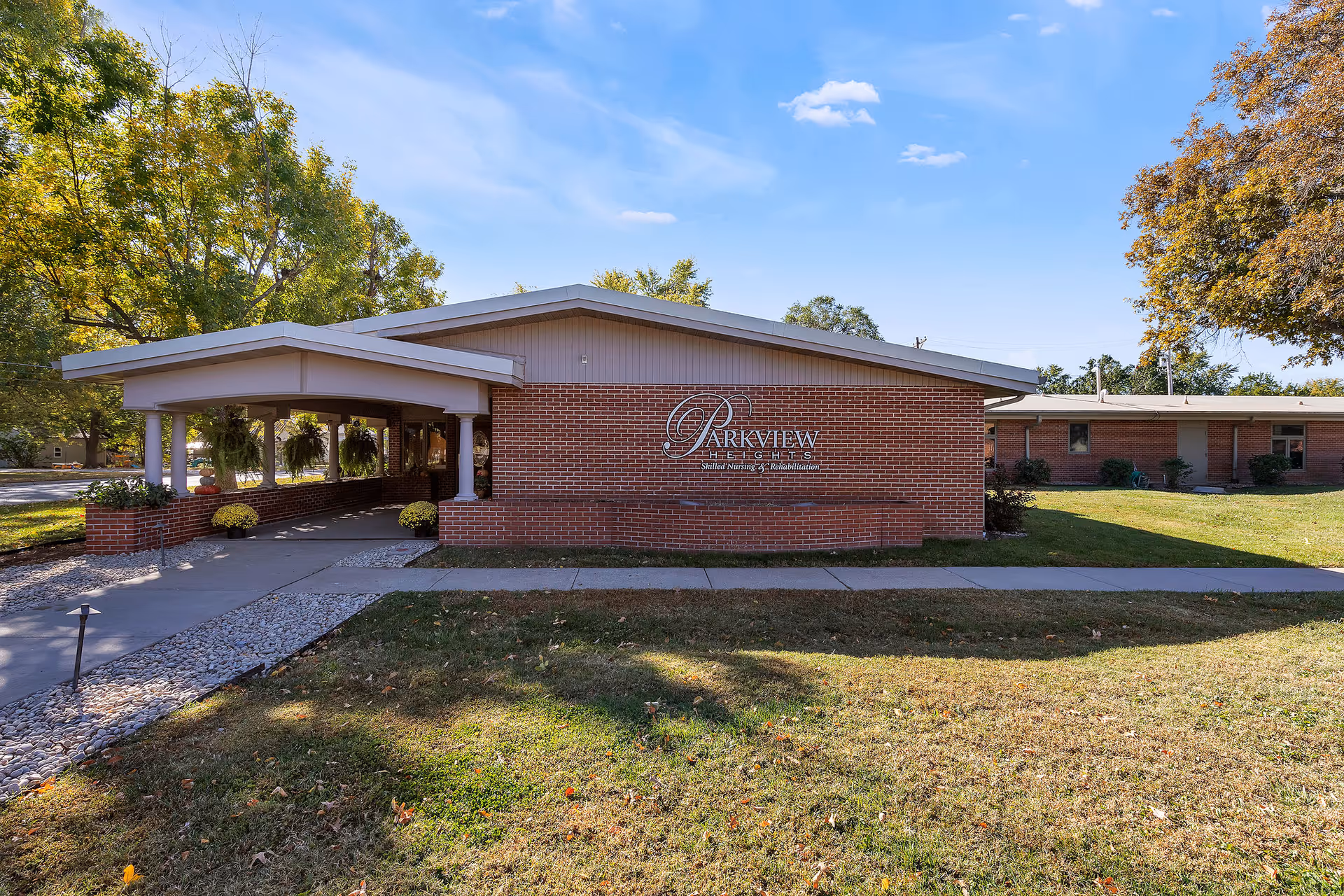 Exterior view of Parkview Heights Care Center, a single-story brick building with a covered entrance and surrounding trees under a clear blue sky.