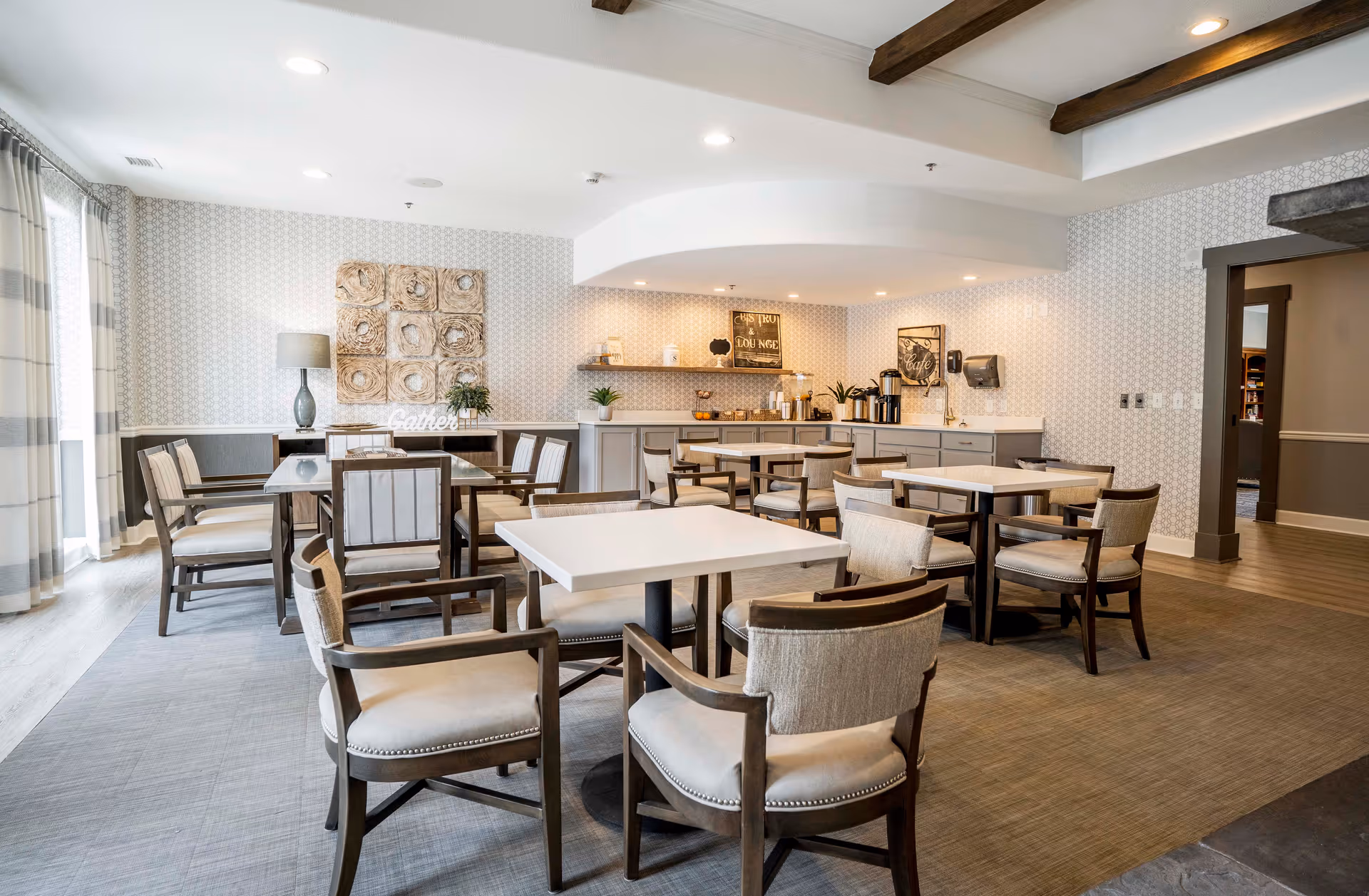 A bright and spacious dining room in an assisted living facility featuring multiple square and rectangular tables surrounded by cushioned wooden chairs. The room has light-colored walls with patterned wallpaper, a large window with sheer curtains allowing natural light, and decorative wall art. In the background, there is a counter area with coffee dispensers, cups, and small plants, creating a welcoming atmosphere.