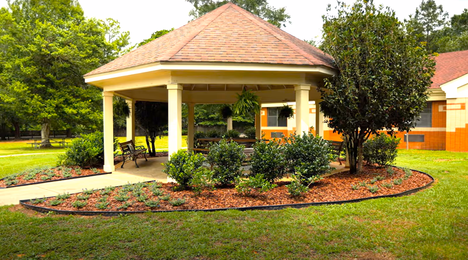 A gazebo with a red shingled roof and cream-colored pillars surrounded by landscaped bushes and mulch in a grassy outdoor area. There are benches inside the gazebo and hanging plants. In the background, part of a brick building is visible along with trees and picnic tables.