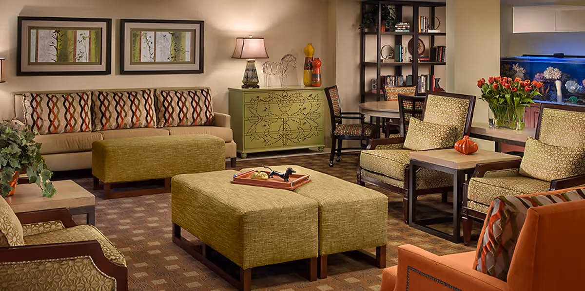 A cozy senior living common area with a beige sofa adorned with patterned cushions, two green upholstered ottomans, and several armchairs with patterned cushions. There is a green decorative cabinet with a lamp and decorative items on top, a round table with chairs, a bookshelf filled with books, and a large aquarium with coral decorations. The room is warmly lit and decorated with framed artwork and a vase of red flowers.