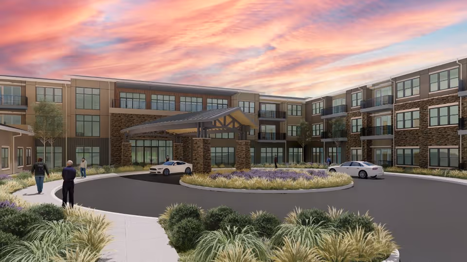 Exterior view of a modern three-story senior living facility with a covered entrance driveway, landscaped roundabout with greenery and purple flowers, several people walking on the sidewalk, and two white cars parked near the entrance under a colorful sunset sky.