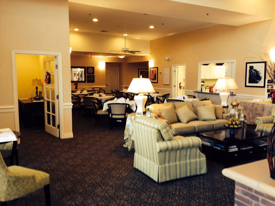 Interior view of a senior living facility showing a cozy common area with beige sofas, armchairs, and a coffee table with decorative items. In the background, there is a dining area with tables and chairs, soft lighting, and framed artwork on the walls.