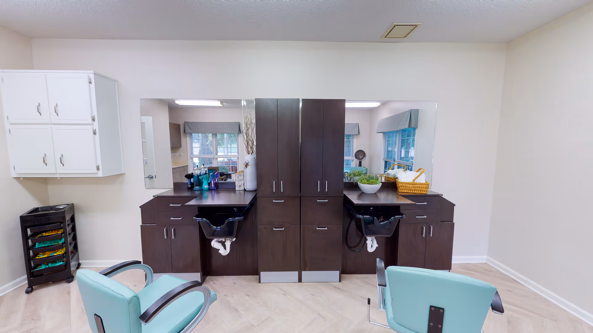 Interior salon area with two teal styling chairs facing a dark wood vanity with sinks and a large mirror.