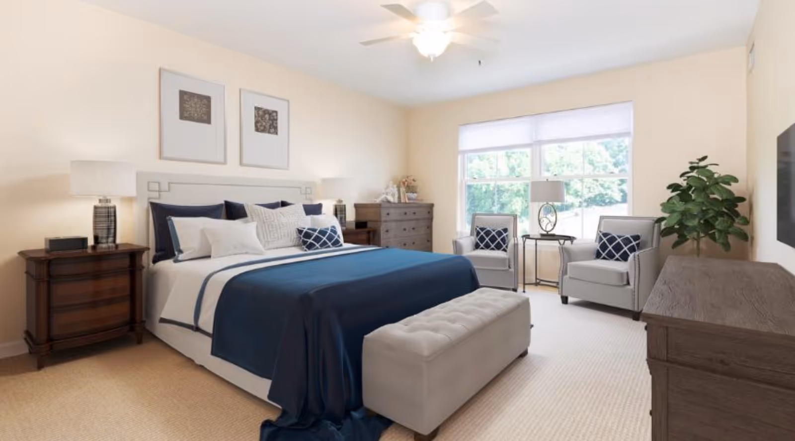 A bright and spacious bedroom with a large bed dressed in white and navy blue bedding, two framed artworks above the headboard, two wooden nightstands with lamps, a cushioned bench at the foot of the bed, two gray armchairs with patterned pillows near a large window, a wooden dresser, a flat-screen TV mounted on the wall, and a potted plant in the corner.