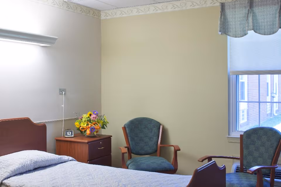 A simple bedroom in a senior living facility with a bed covered in a light blue quilt, a wooden nightstand with a colorful flower arrangement and a small clock, two green upholstered chairs, and a window with a blue valance showing an exterior brick wall.
