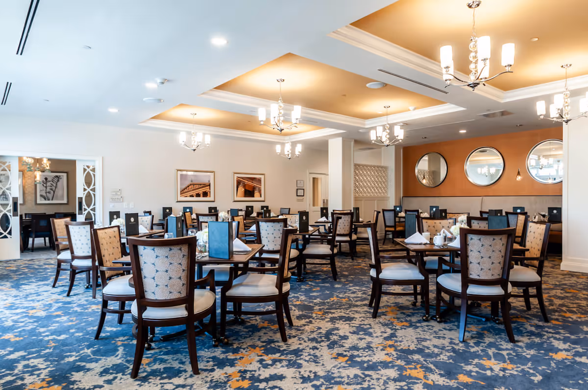 A spacious dining room with multiple wooden tables and upholstered chairs arranged neatly. The room features a patterned blue and orange carpet, white walls adorned with framed pictures and round mirrors, and elegant chandeliers hanging from a recessed ceiling with warm lighting.