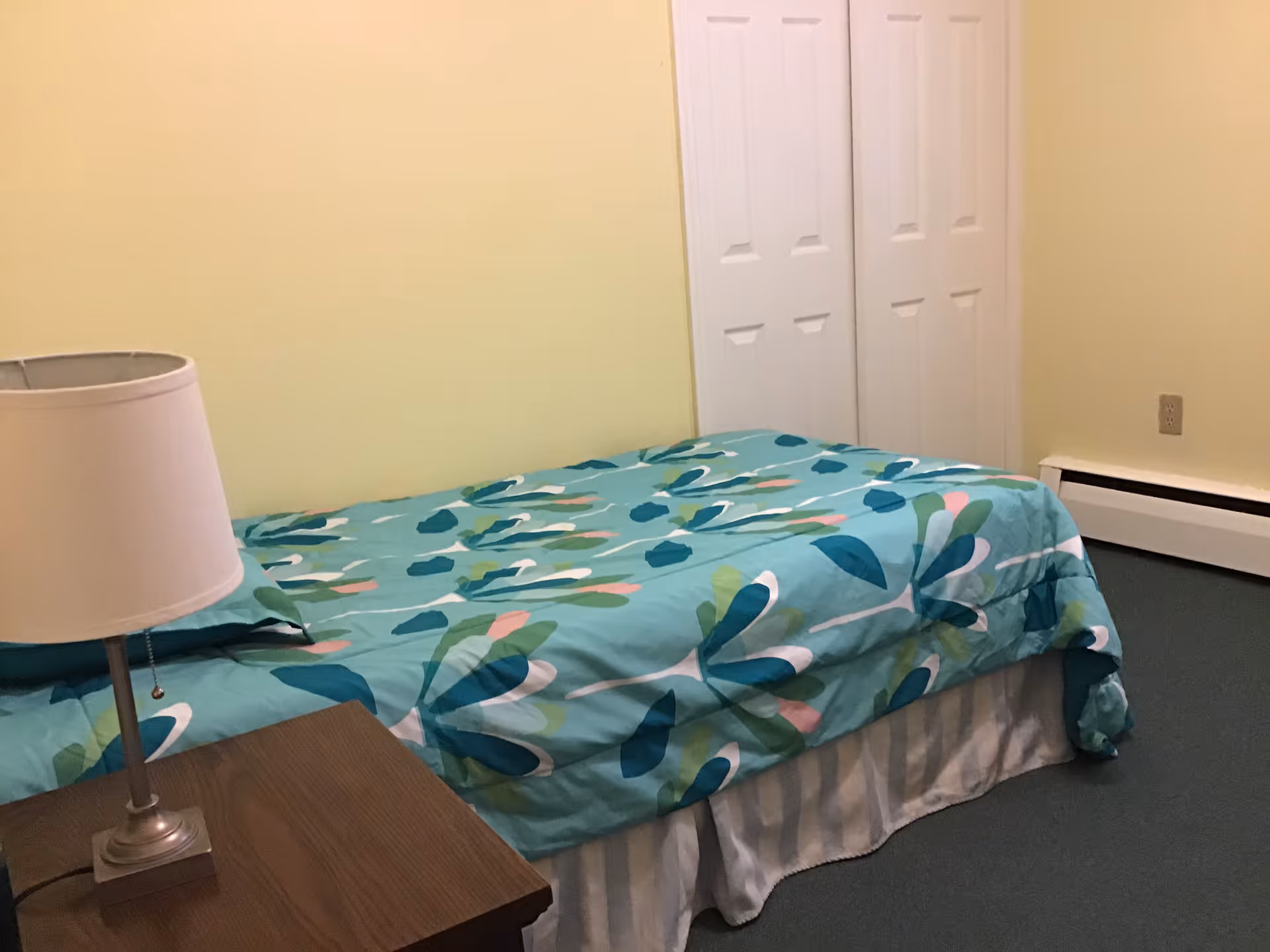 A simple bedroom with a single bed covered in a blue and green patterned bedspread. There is a wooden nightstand with a lamp on it next to the bed. The walls are painted light yellow, and there is a white double-door closet in the background.