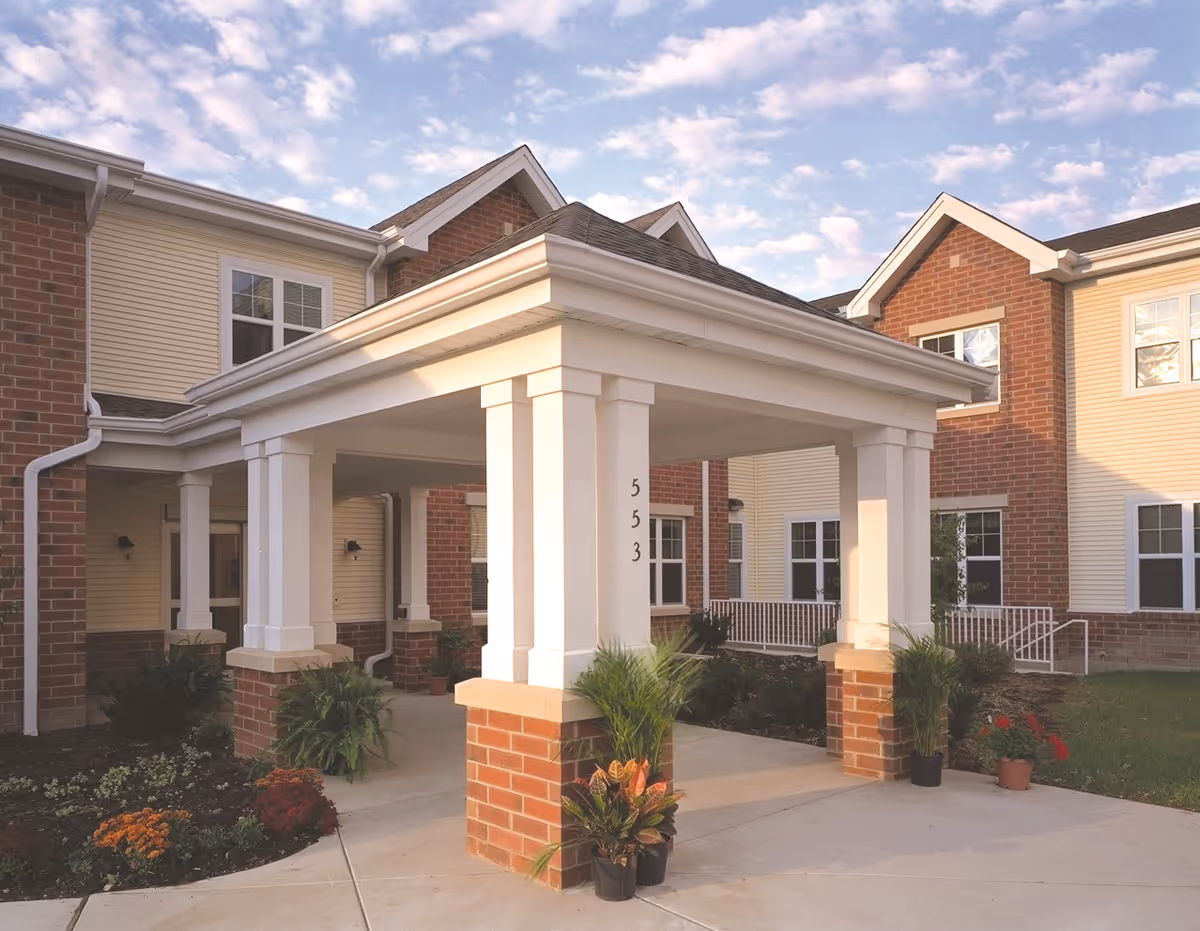 Entrance of a senior living facility named Addolorata Villa with a covered porch supported by white columns with brick bases, surrounded by plants and flowers, under a partly cloudy sky.