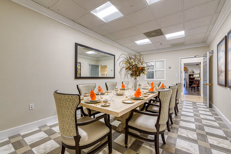A well-lit dining room with a long rectangular table set for eight people. The table is decorated with a large floral centerpiece and each place setting includes a plate, cup, glass, and an orange folded napkin. The room has patterned chairs, a large mirror on one wall, two framed paintings on another, and a checkered tile floor. A doorway at the far end leads to another room.