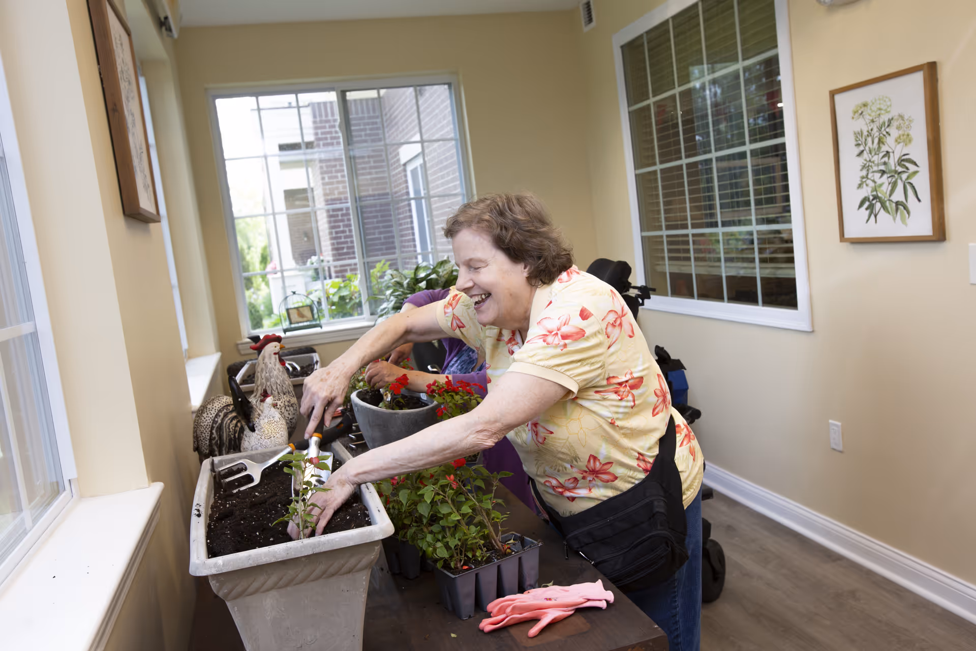 An elderly woman with short brown hair wearing a yellow floral shirt is happily planting flowers in a rectangular planter indoors. She is using a small gardening tool and is surrounded by other potted plants and gardening gloves on a wooden table. The room has large windows with white frames, beige walls, and framed botanical artwork on the wall.