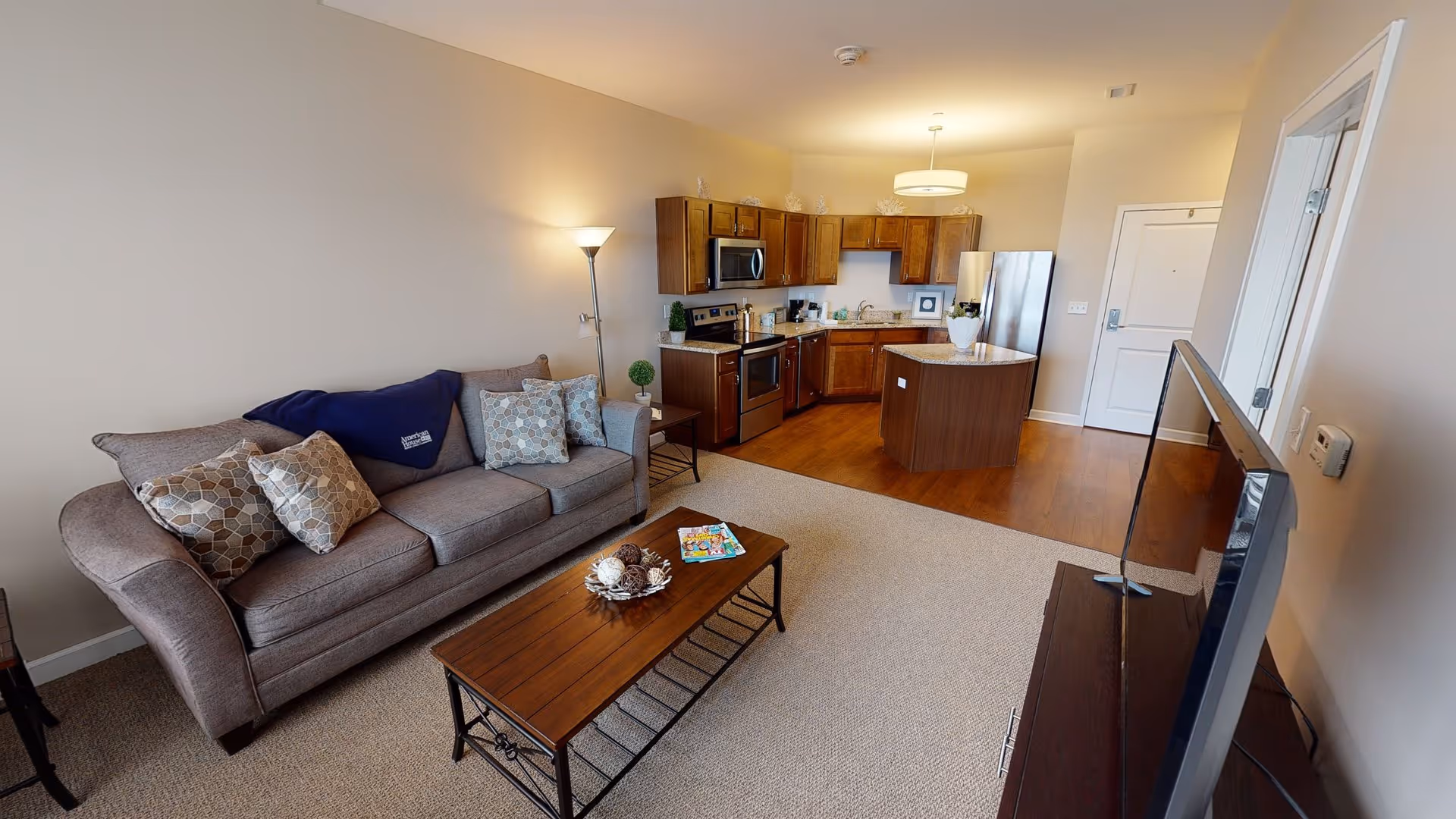 A cozy living room and kitchen area in an apartment at American House Lakeshore. The living room features a gray sofa with patterned cushions and a blue throw blanket, a wooden coffee table with decorative items and magazines, and a floor lamp. The kitchen has wooden cabinets, stainless steel appliances including a microwave, stove, dishwasher, and refrigerator, and a small island with a plant on top. The floor transitions from carpet in the living area to wood in the kitchen.