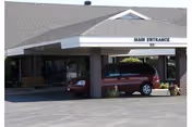 Exterior view of the main entrance of a senior living facility with a covered drop-off area, a red minivan parked under the canopy, and a bench near the entrance.