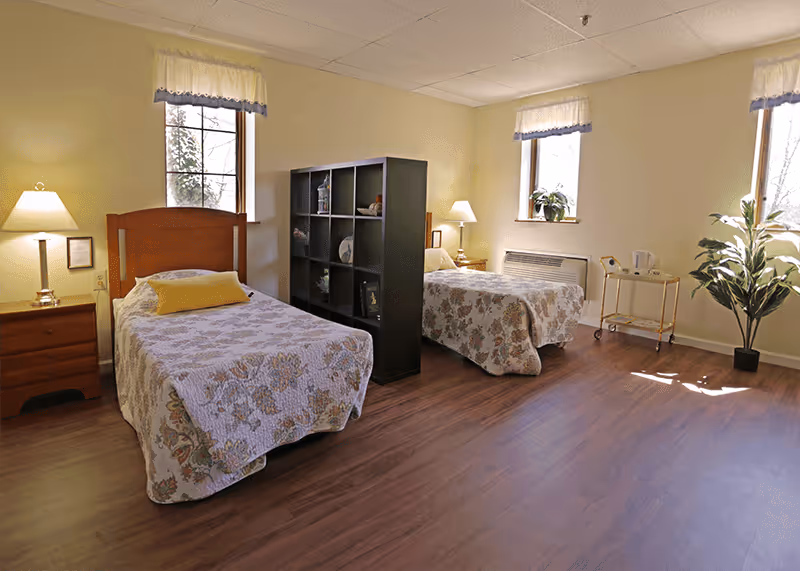 A bright bedroom with two single beds separated by a black shelving unit. Each bed has a floral bedspread and a pillow. There are two windows with white curtains, a wooden nightstand with a lamp, a small table with a kettle, and a potted plant near the window. The floor is wooden, and the walls are painted light yellow.