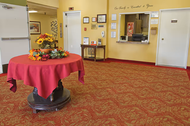 Reception area with a round table covered with a red tablecloth and decorated with a floral arrangement. The walls are painted yellow and there is a reception window with a desk and chair behind it. Various framed certificates and notices are displayed on the wall around the reception window. The carpet has a red and gold ornate pattern. There are two white doors on either side of the reception window, and a small table with brochures and decorations is placed against the wall.