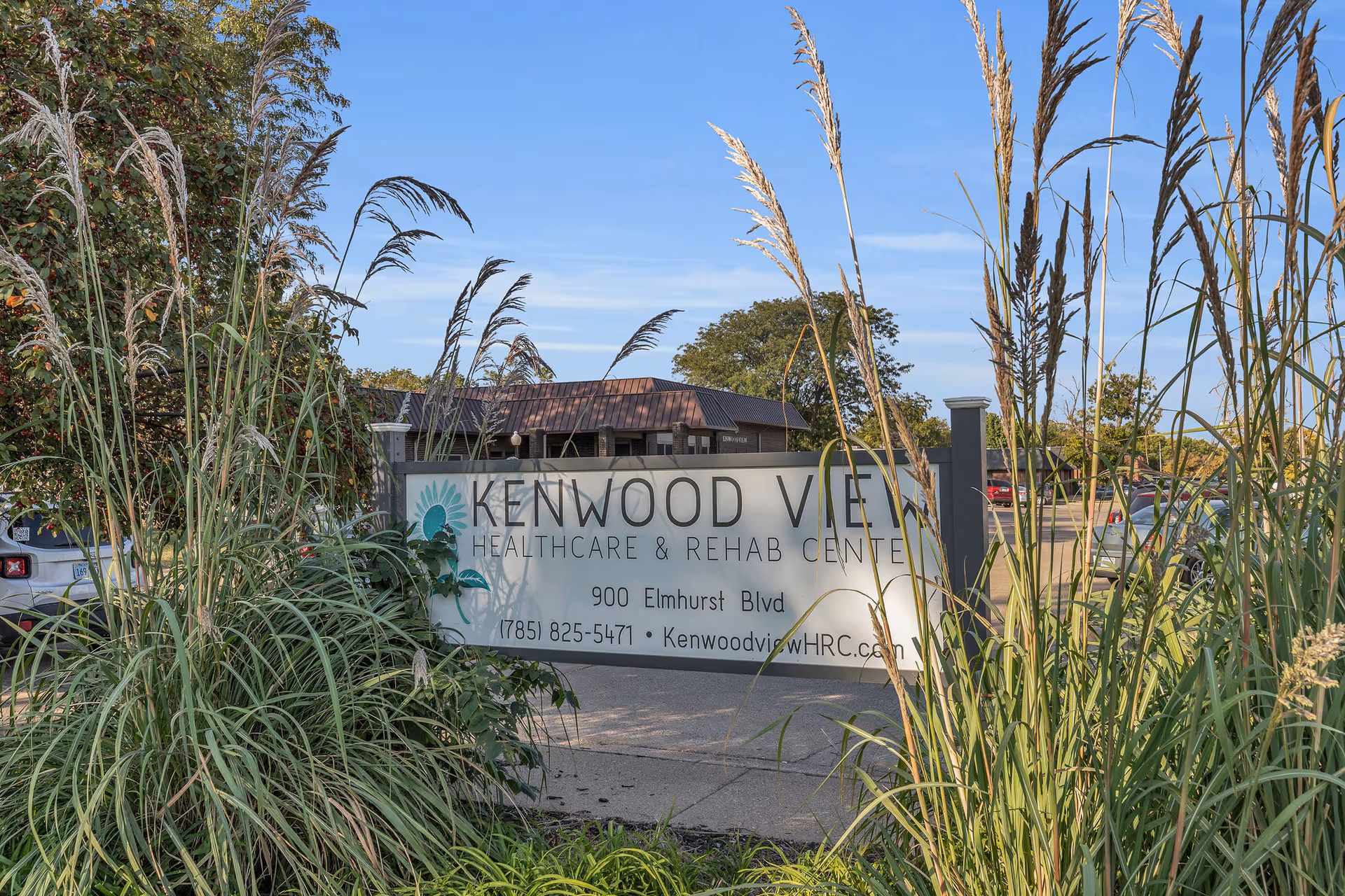 Outdoor view of the entrance sign for Kenwood View Healthcare & Rehab Center, partially obscured by tall ornamental grasses, with a parking lot and building visible in the background under a clear blue sky.