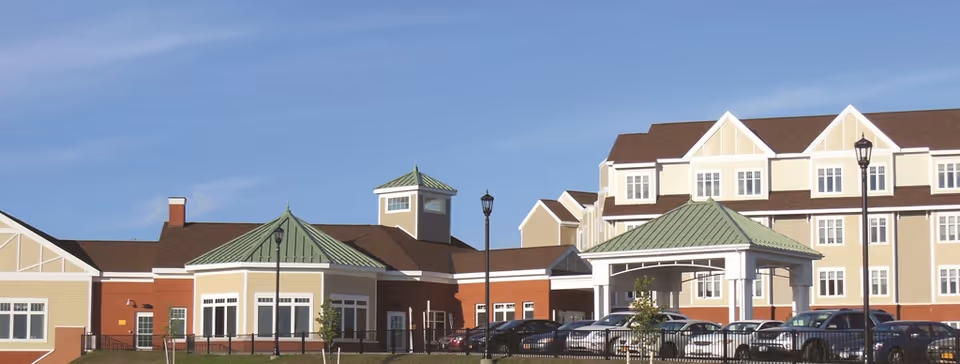 Exterior view of Samaritan Summit Village, a multi-story senior living facility with beige and brick walls, green metal roofs, and a covered entrance. Several cars are parked in front of the building under a clear blue sky.