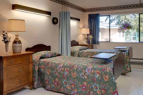 Two single beds with floral bedding, nightstands, lamps, and a windowed wall in a shared room.