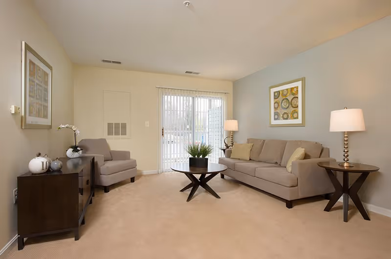 A living room with a beige sofa and matching armchair, two wooden side tables with lamps, a round wooden coffee table with a plant, a wooden cabinet with decorative items, and a large window with vertical blinds letting in natural light.