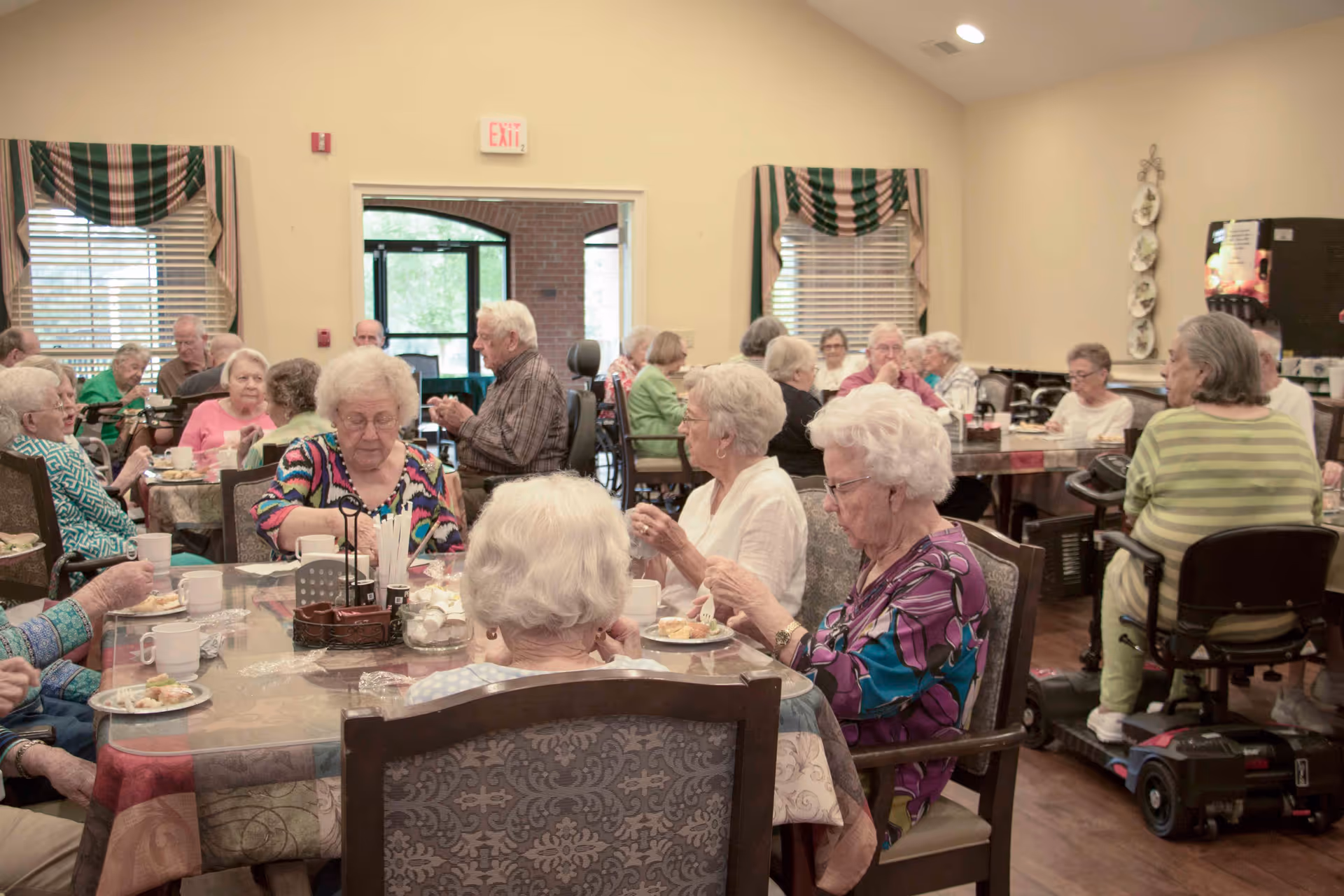 A group of elderly people sitting at multiple tables in a dining room, eating and socializing. The room has large windows with striped valances, and there is a beverage machine in the background. Some individuals are using mobility scooters.