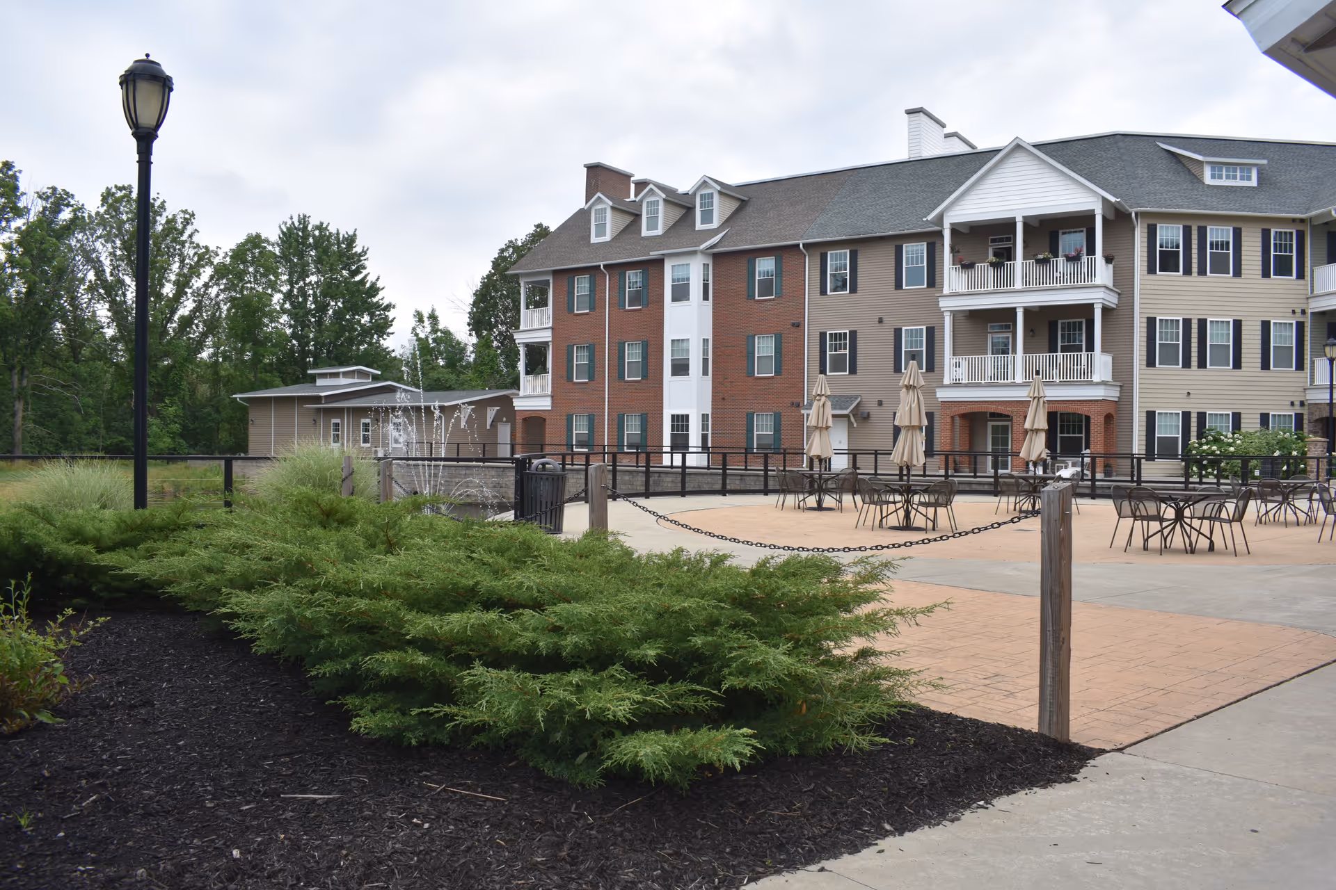 Outdoor patio area with tables and chairs with closed umbrellas in front of a multi-story residential building. There are green shrubs and a lamppost in the foreground, and trees in the background under a cloudy sky.