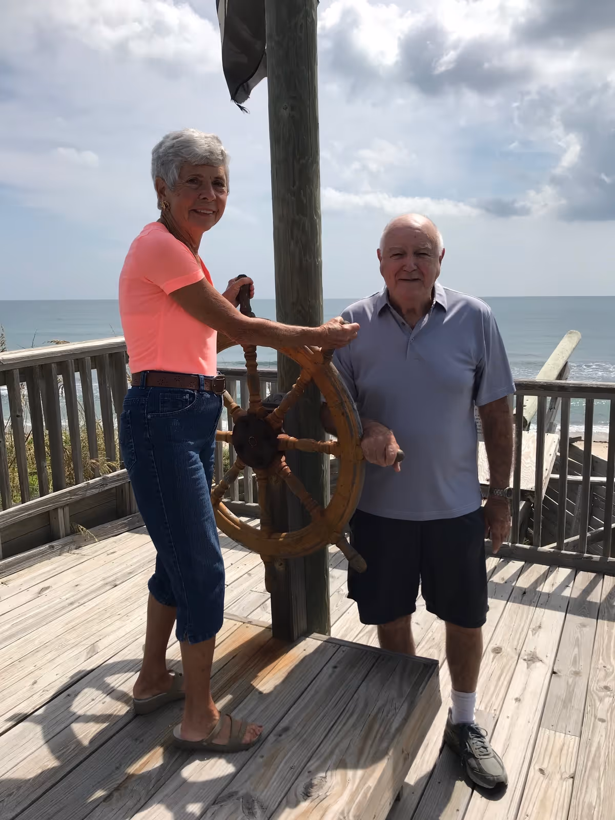 An elderly woman and man standing on a wooden deck by the ocean. The woman is holding a large wooden ship's wheel mounted on a post, and the man stands beside her. The sky is partly cloudy and the ocean is visible in the background.