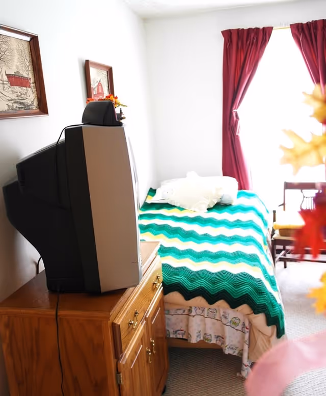 A small bedroom with a single bed covered by a green, yellow, and white zigzag patterned blanket. There is a wooden cabinet with an old-style CRT television on top. The room has white walls with two framed pictures hanging. A window with red curtains allows bright light to enter the room. A wooden chair is placed near the window.