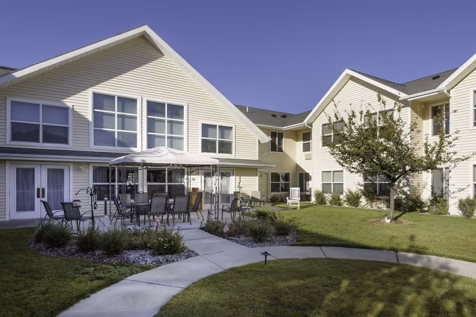 Courtyard with patio seating and a canopy surrounded by a two-story beige senior living building and landscaped lawn.