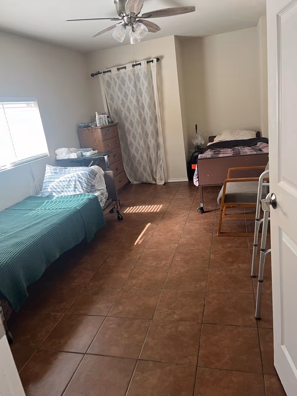 A simple bedroom with two single beds, one on each side of the room. The bed on the left has a green blanket and striped pillow, while the bed on the right has a pink and black blanket. There is a wooden dresser with various items on top near the back wall, a chair next to the right bed, a walker near the door, and a ceiling fan with lights above. The floor is tiled, and a window on the left wall lets in natural light.