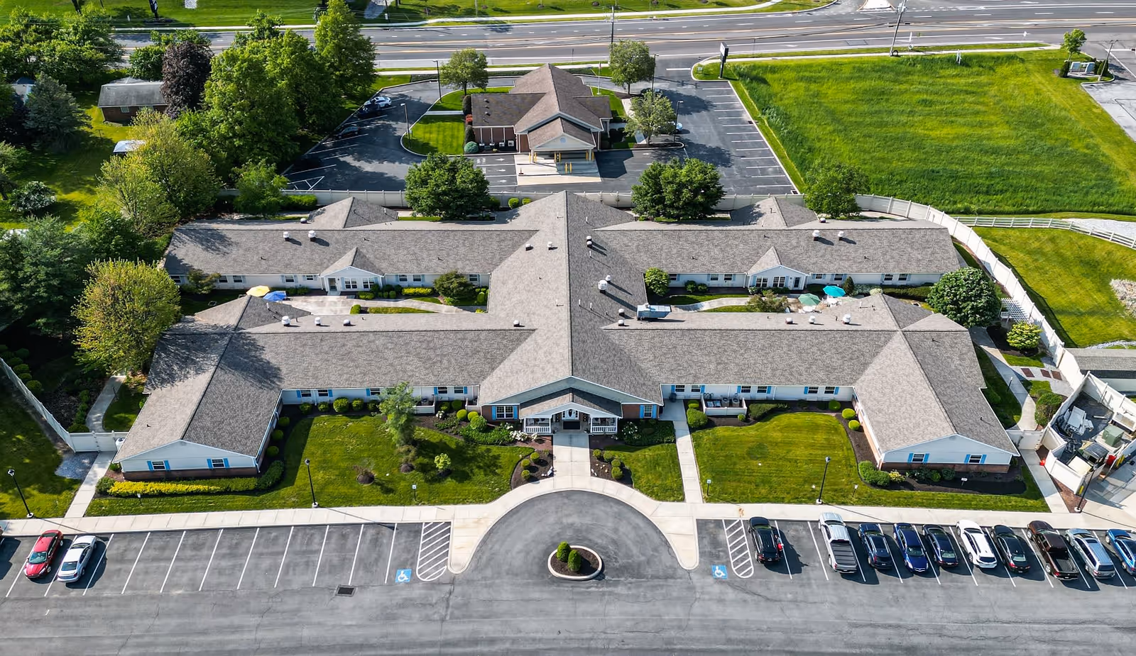 Aerial view of Arden Courts - ProMedica Memory Care Community (Susquehanna), showing a large single-story building with a central entrance, surrounded by well-maintained green lawns, trees, and parking lots with several cars parked. The building has a gray roof and white exterior walls, with sidewalks leading to the entrance and around the property. A road runs behind the facility.