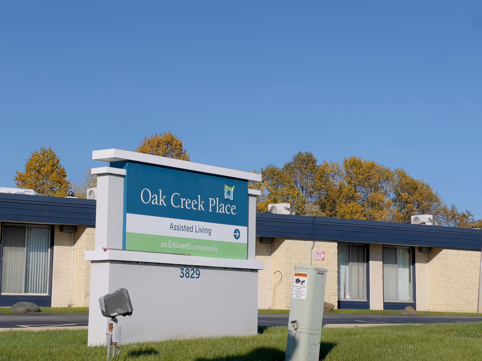 Outdoor view of the Oak Creek Place assisted living facility sign with the building and trees in the background under a clear blue sky.