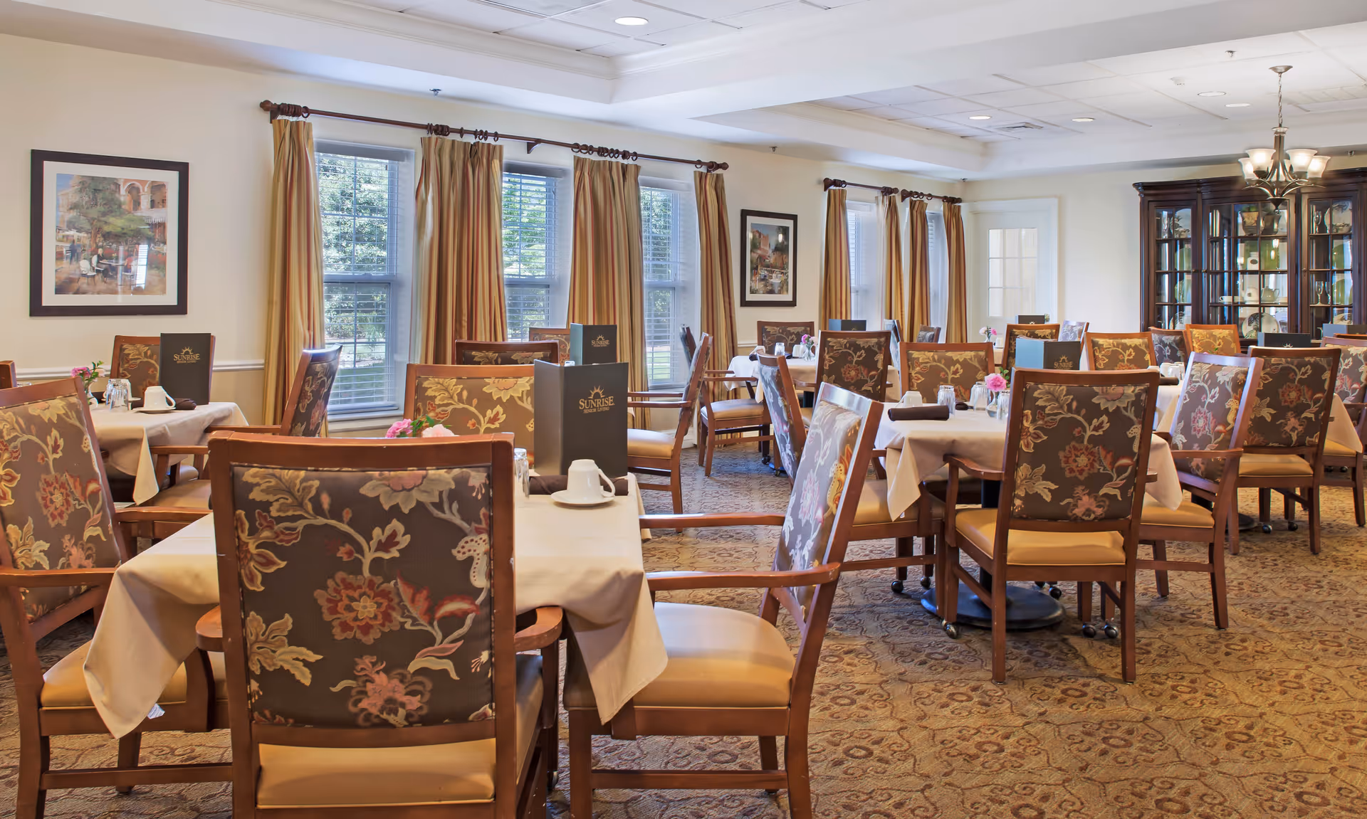 A well-lit dining room with multiple tables covered in white tablecloths, each surrounded by wooden chairs with floral-patterned upholstery. Menus labeled 'Sunrise Senior Living' are placed on several tables. The room has large windows with beige curtains, framed artwork on the walls, a chandelier, and a glass-fronted cabinet displaying decorative items.