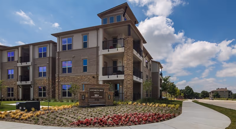 Three-story senior living building with brick exterior, balconies, and a curved landscaped walkway at the front.