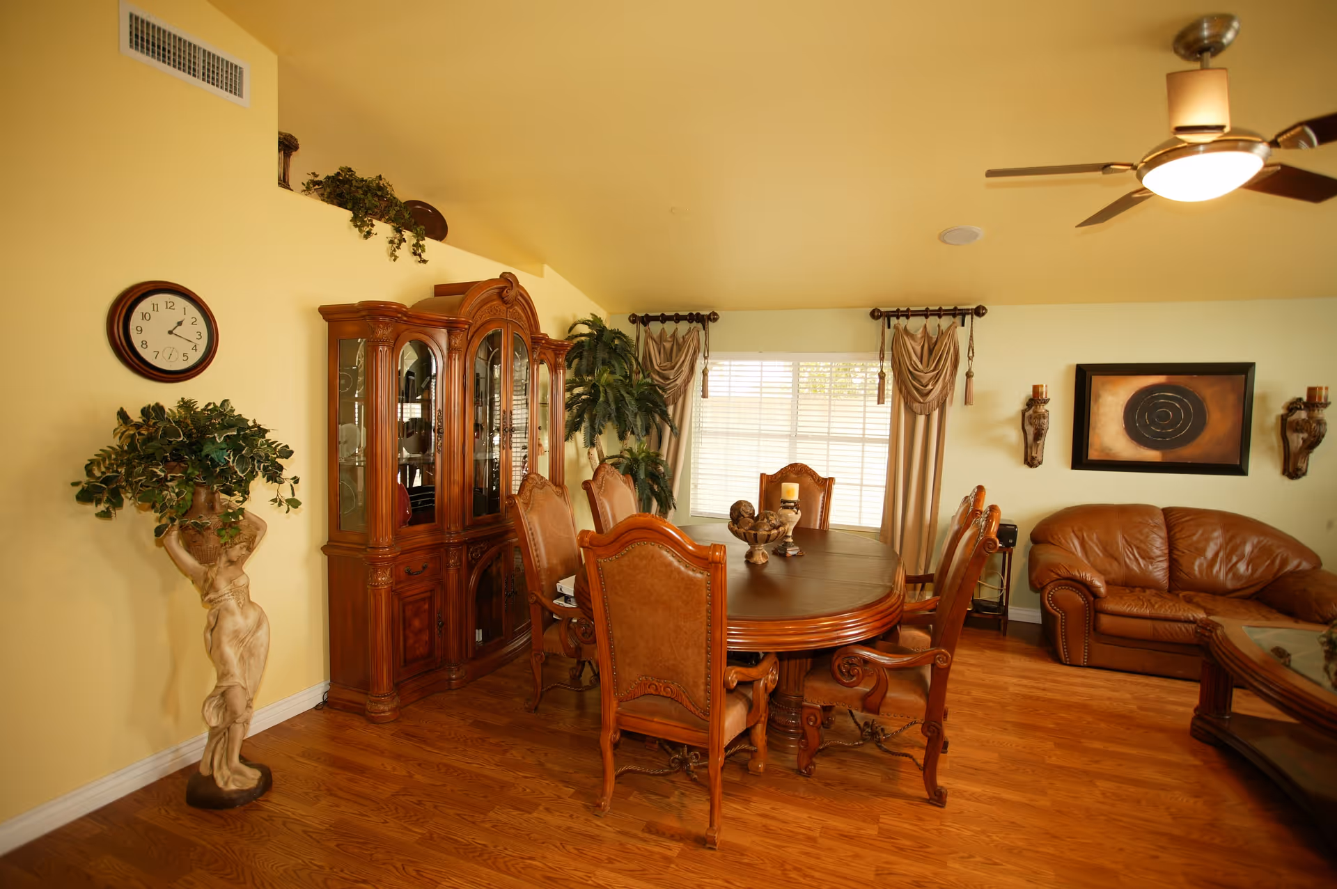 A warmly lit dining and living area with a wooden dining table surrounded by six upholstered chairs. A large wooden china cabinet stands against a yellow wall with a round clock above it. There is a decorative statue holding a plant on the left side and a large window with curtains behind the dining table. To the right, a brown leather couch and a coffee table are visible under a ceiling fan with a light. The room has wooden flooring and soft yellow walls.