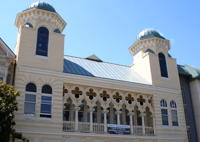 Exterior view of a historic-style building with two towers featuring domed roofs and arched windows. The building has decorative architectural details and a sign that reads 'Kokoro Assisted Living NOW ACCEPTING RESIDENCY APPLICATIONS'. The sky is clear and blue.
