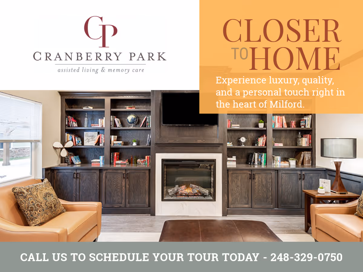 A cozy living room area with two tan leather armchairs, a dark wooden built-in bookshelf filled with books and decorative items, a fireplace in the center, and a flat-screen TV mounted above the fireplace. There is a window with blinds on the left side and a table lamp on a side table to the right.