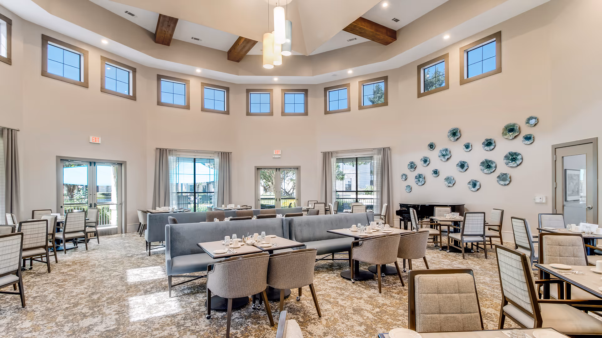A spacious dining room with multiple tables and chairs arranged neatly. The room features high ceilings with wooden beams and multiple small windows near the ceiling allowing natural light to enter. There are large windows and glass doors on the walls, with light gray curtains. The walls are decorated with blue flower-shaped wall art, and there is a black grand piano in the corner. The tables are set with white plates, cups, and silverware.