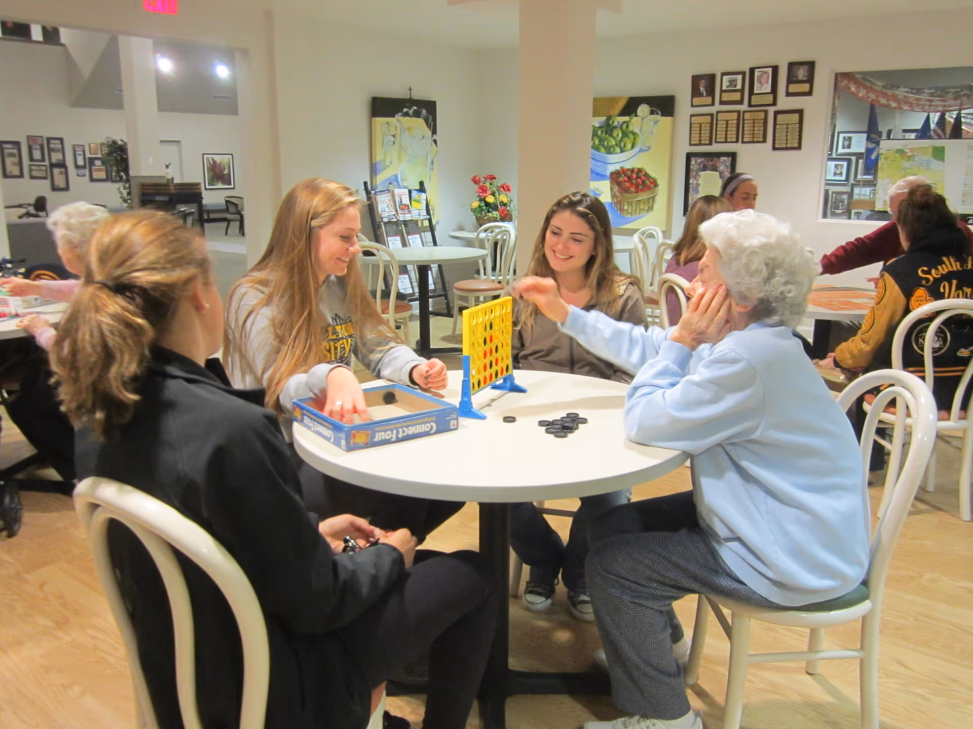 A group of people, including an elderly woman and younger adults, sitting around a round table playing the board game Connect Four in a well-lit common area with chairs and tables. The background shows other people seated and wall decorations including paintings and plaques.