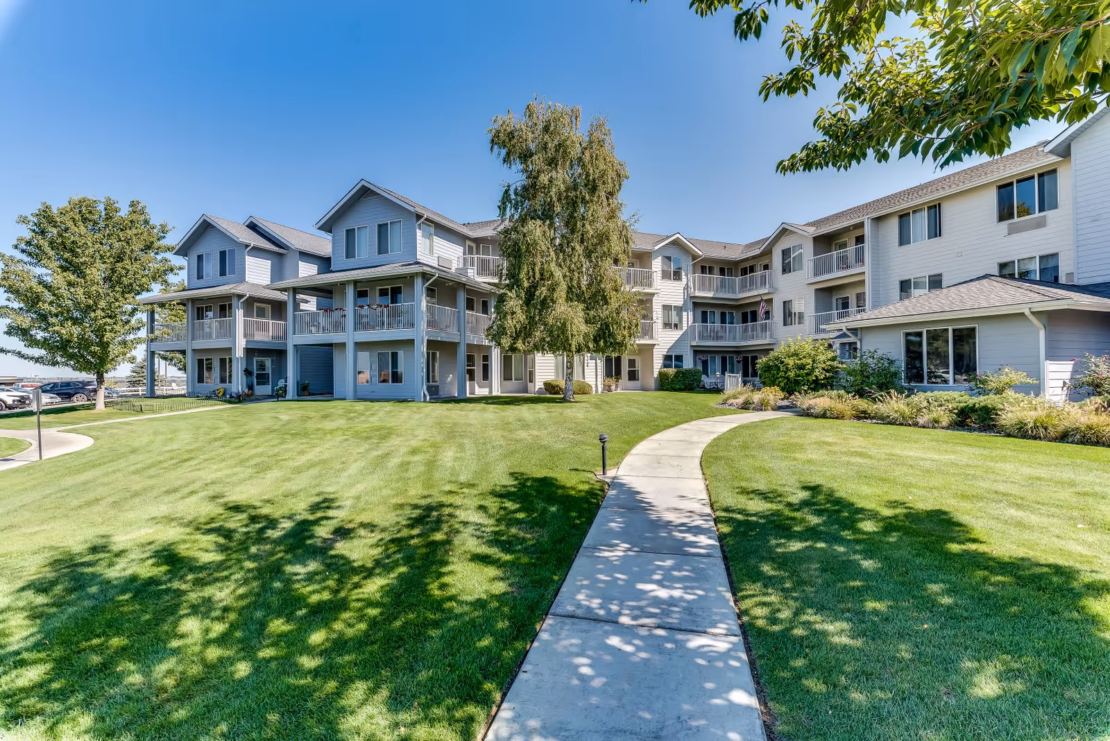 Exterior view of a senior living facility with a well-maintained lawn, a concrete walkway leading to the entrance, and a three-story building with balconies and large windows under a clear blue sky.