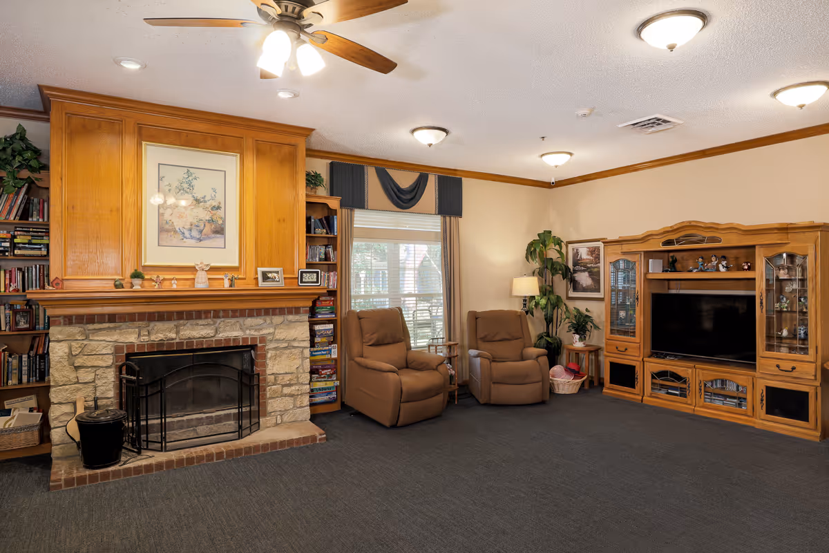 A cozy living room with a stone fireplace and wooden mantel decorated with small figurines and a framed floral painting above it. To the left of the fireplace are built-in wooden bookshelves filled with books. Two brown recliner chairs are positioned near a window with curtains and a valance. On the right side, there is a large wooden entertainment center with a flat-screen TV and decorative items inside glass cabinets. The room has a ceiling fan with lights and several ceiling-mounted light fixtures, beige walls with wooden trim, and a dark carpeted floor.