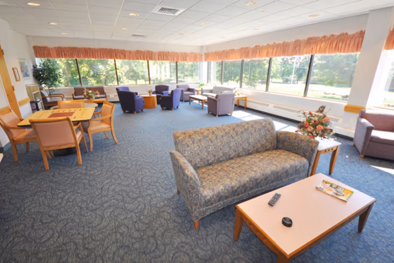 Sunlit communal lounge with sofas, armchairs, tables, and large windows overlooking greenery.