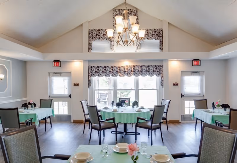 A bright dining room with several tables covered in light green tablecloths, each set with plates, glasses, and napkins. The room has high vaulted ceilings with a chandelier, large windows with floral valances, and two exit doors on either side of the windows.