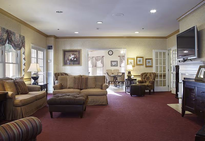 A cozy living room area with beige sofas, a cushioned ottoman, armchairs, and side tables with lamps. The room has a red carpet, framed artwork on the walls, and a flat-screen TV mounted above a fireplace. Large windows with patterned valances allow natural light to enter. In the background, there is a dining area with a round table and chairs.