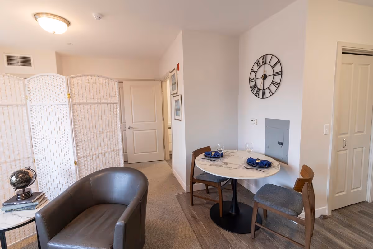 A small dining area with a round marble table set for two with blue napkins and wine glasses, two wooden chairs with gray cushions, a large wall clock above the table, a gray armchair next to a small round side table with a decorative globe and books, and a white folding room divider in the background near a closed door.