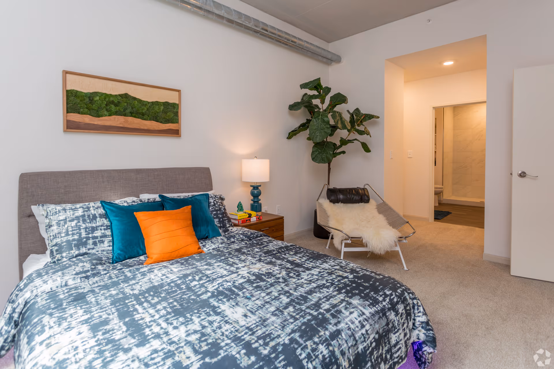 A modern bedroom with a gray upholstered bed featuring blue and orange pillows and a patterned blue and white comforter. Next to the bed is a wooden nightstand with a blue lamp and some books. A large green leafy plant is placed beside a white chair with a fur throw. The room has beige carpet and white walls, with a framed artwork above the bed. An open doorway leads to a bathroom with a visible toilet and shower area.