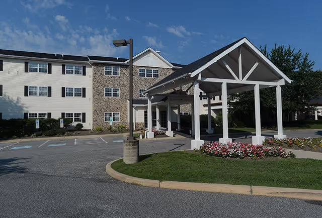 Exterior view of Glen Mills Senior Living facility showing a three-story building with a combination of stone and siding facade, a covered entrance supported by white pillars, landscaped flower beds, and a parking area with handicap spaces under a clear blue sky.