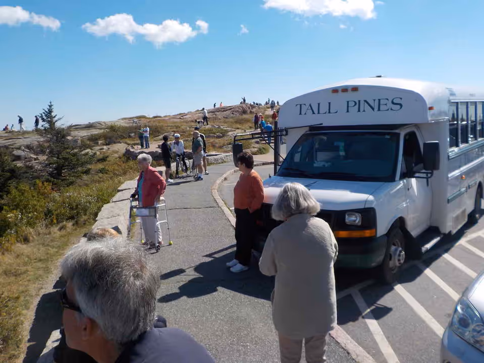 Seniors standing near a Tall Pines shuttle bus at a scenic outdoor overlook with rocky hills and blue sky.